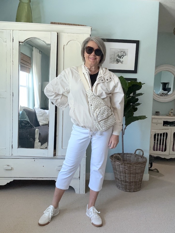 Woman over 50 styling white capri pants with a black t-shirt, cream crossbody bag, and white sneakers for a casual summer outfit.