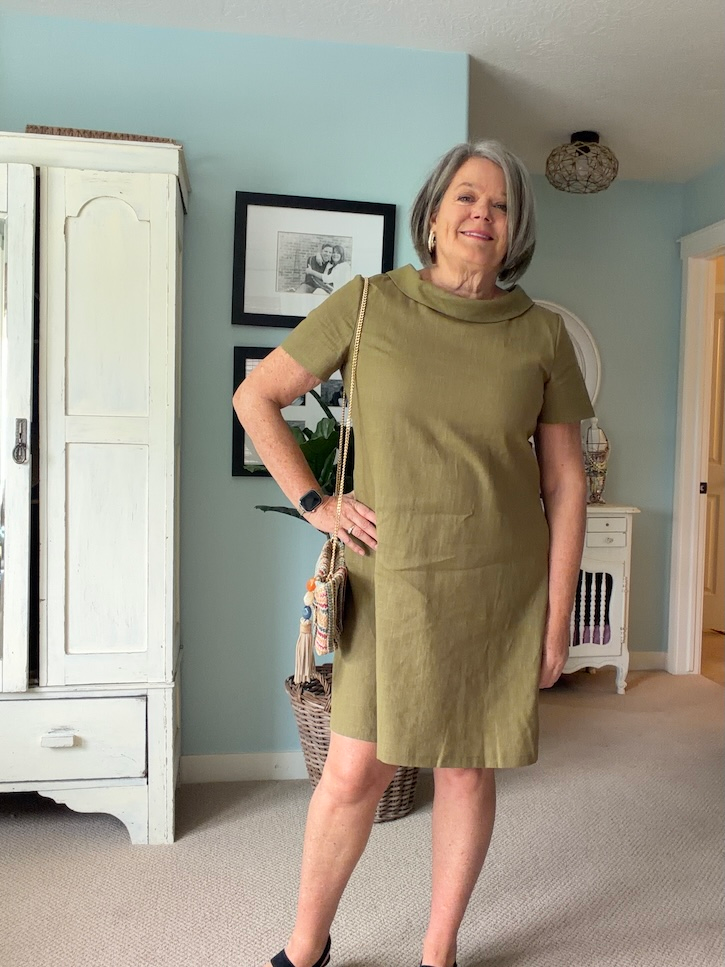 Woman over 50 wearing an olive green LeMuse linen dress with a wide brim sun hat and woven bag