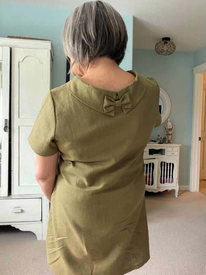 Woman over 50 wearing an olive green LeMuse linen dress with a wide brim sun hat and woven bag