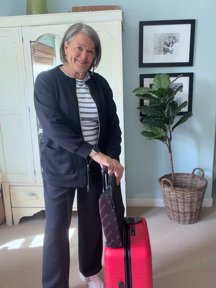 Woman over 50 dressed for a flight to Italy with a red carry-on suitcase and backpack.