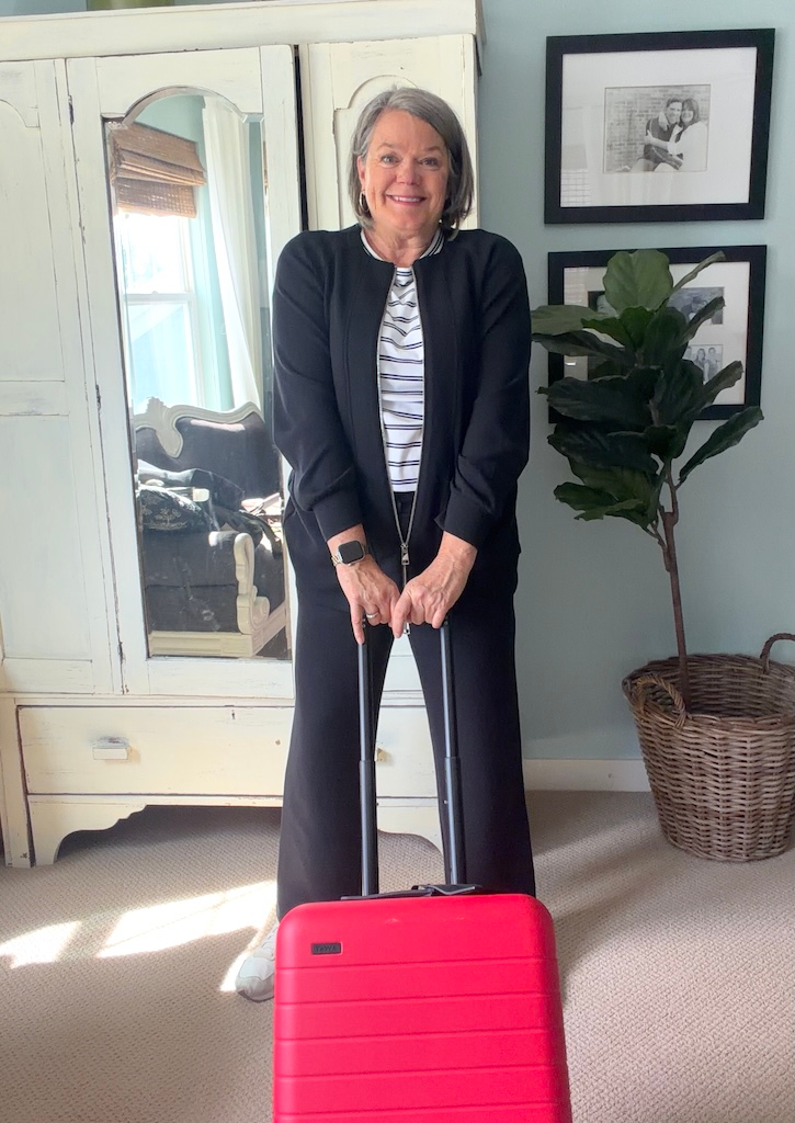 Woman over 50 wearing a black travel outfit with red Away carry-on luggage for a 17-day Italy trip.