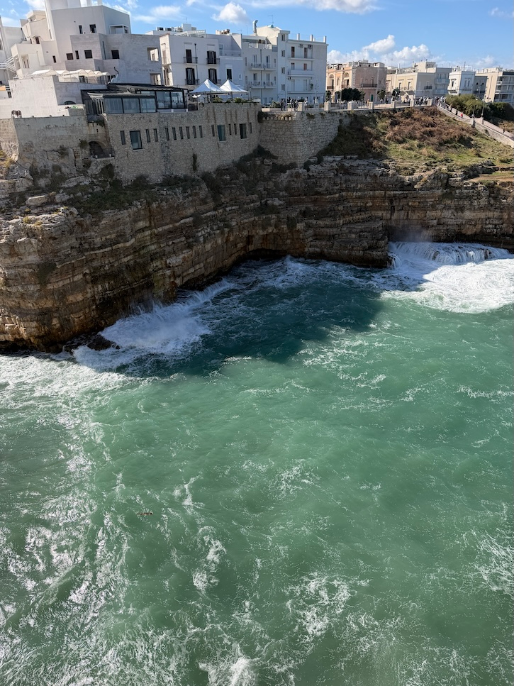 Turquoise sea crashing against rocky cliffs below white buildings along the Puglia coast