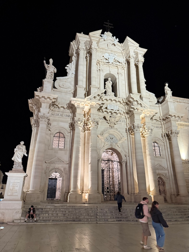 Grand baroque church glowing at night in Italy with people gathered on the steps.