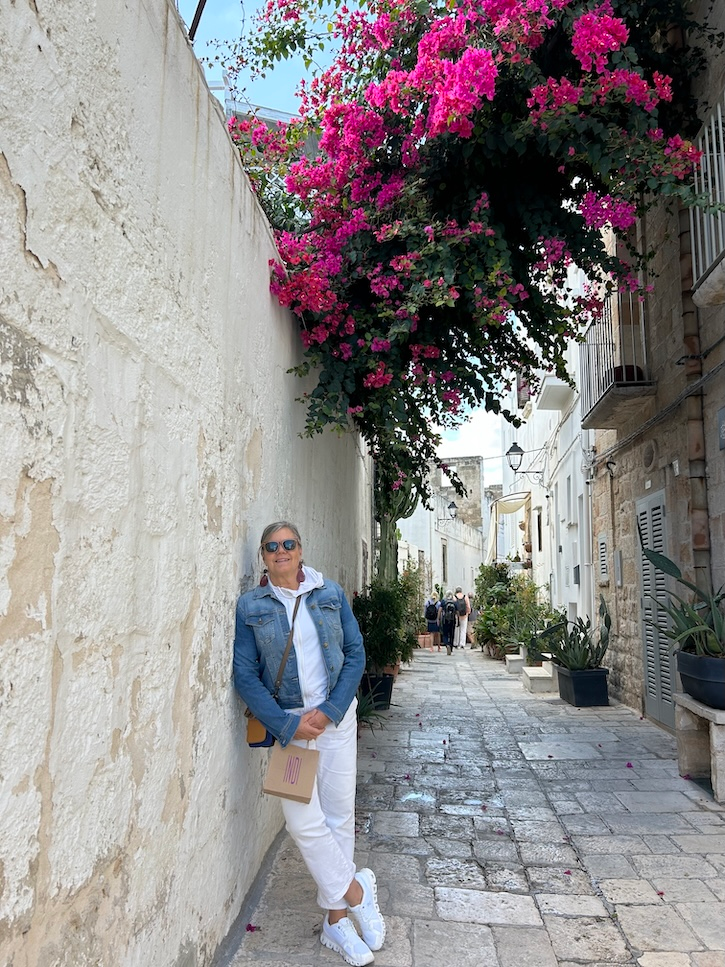 Woman over 50 wearing white pants, a white zip sweatshirt, denim jacket, and white sneakers beneath bright pink bougainvillea in Italy.