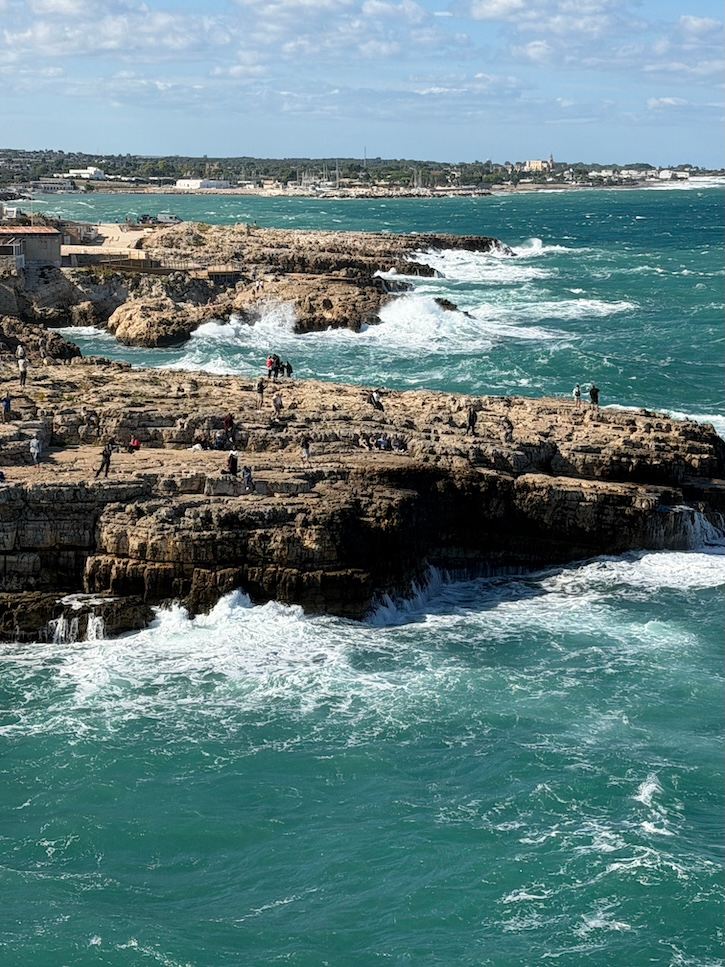 Waves crashing over dramatic rocky coastline in Puglia, Italy under a bright blue sky.