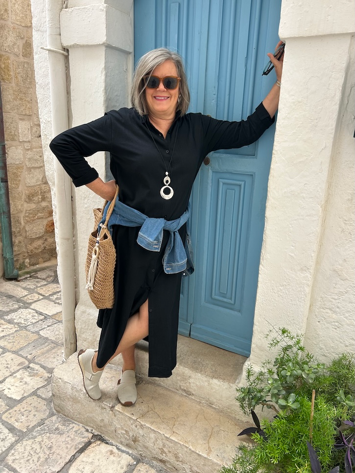 Woman over 50 wearing a black button-down maxi shirt dress with a denim jacket tied at her waist and BØRN sneakers in Bari, Italy.