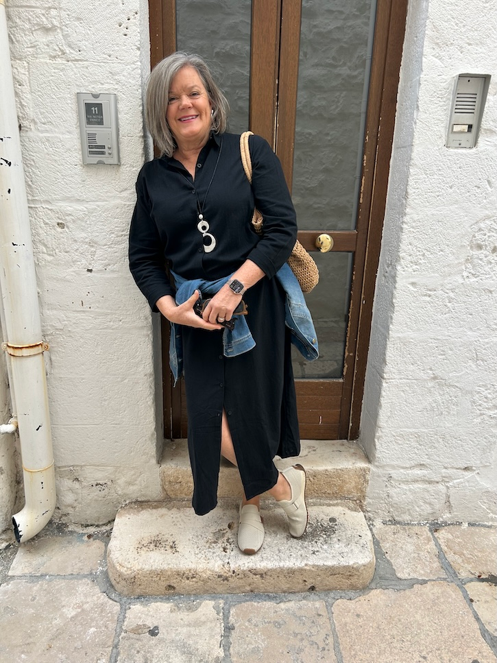 Woman over 50 wearing a black button-down maxi shirt dress with a denim jacket tied at her waist and BØRN sneakers in Bari, Italy.